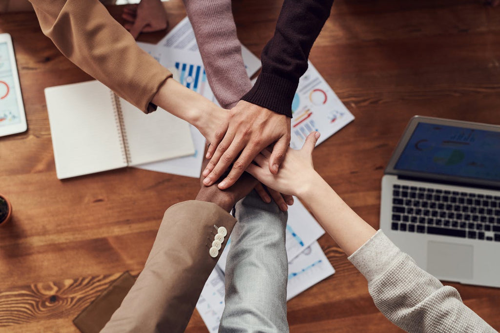 6 hands stacked on top of each other, like a team about to cheer when they leave a huddle. They are standing over a desk with paper and a laptop.