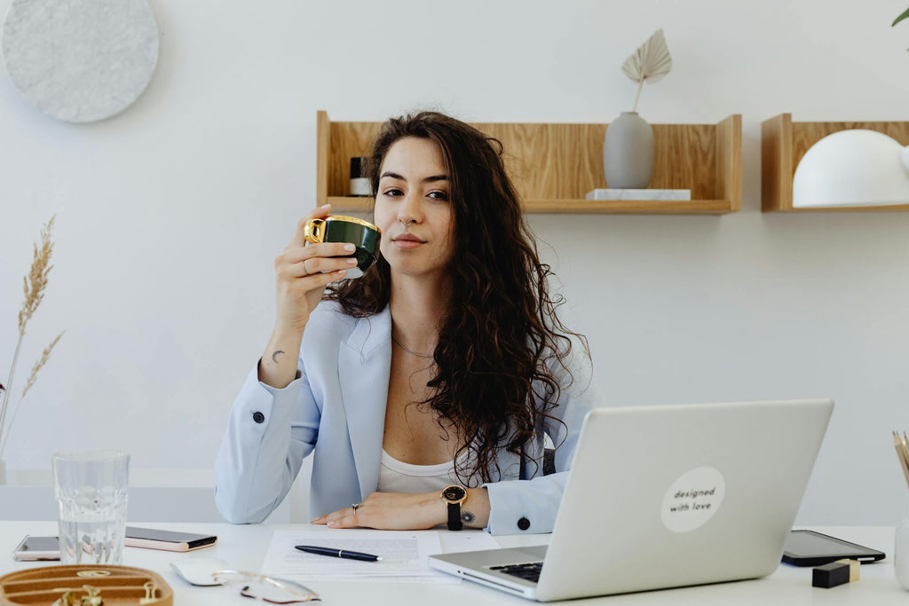 Woman with long brown hair sits at a computer, drinking her coffee while making eye contact with the camera