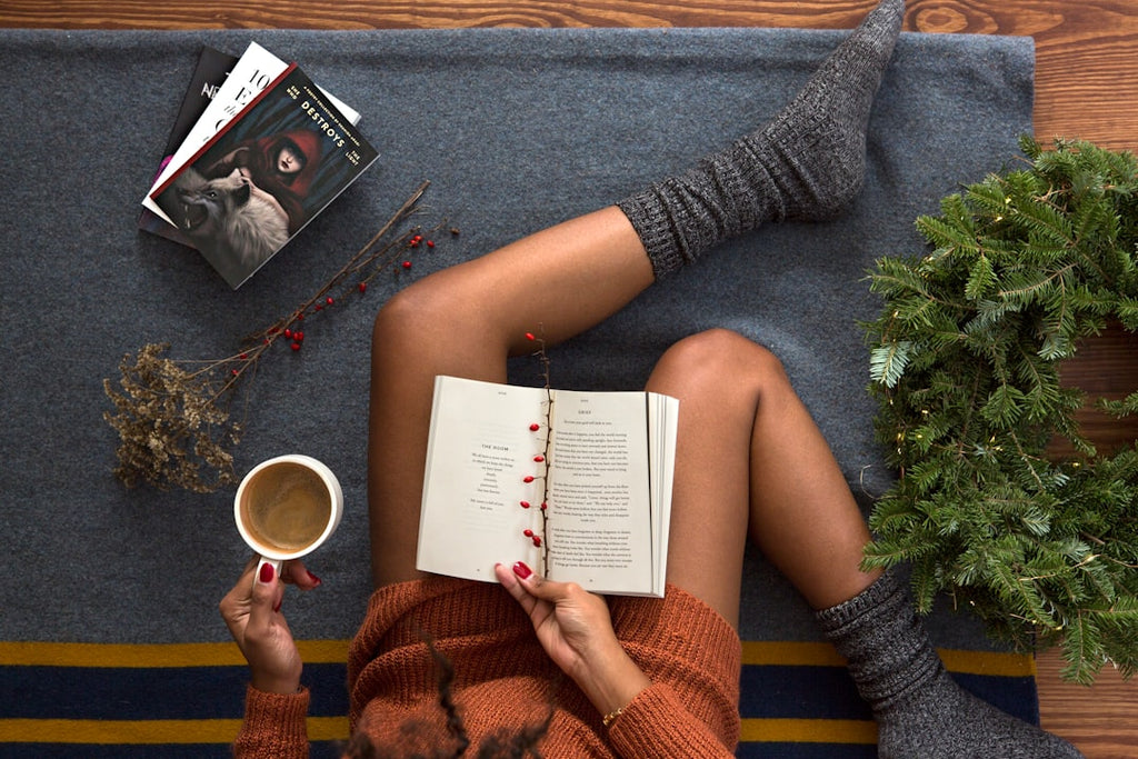 Person relaxing with coffee and book, cozy socks, next to Christmas decor, suggesting gift ideas.