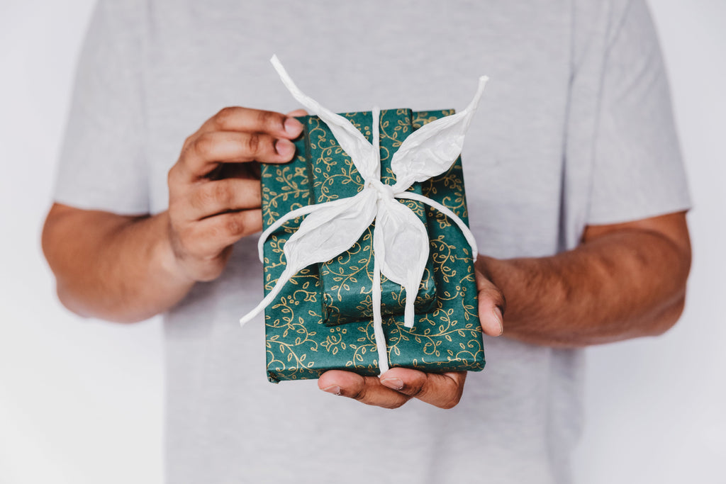 Hands holding a beautifully wrapped present in teal and gold paper with a white ribbon tied like a flower in front of a white shirt.